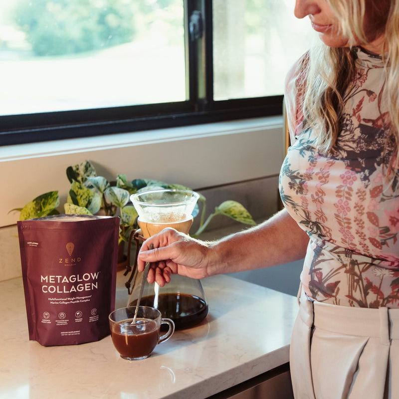 A mother at her kitchen counter, baby on her hip, stirring a scoop of MetaGlow into her coffee