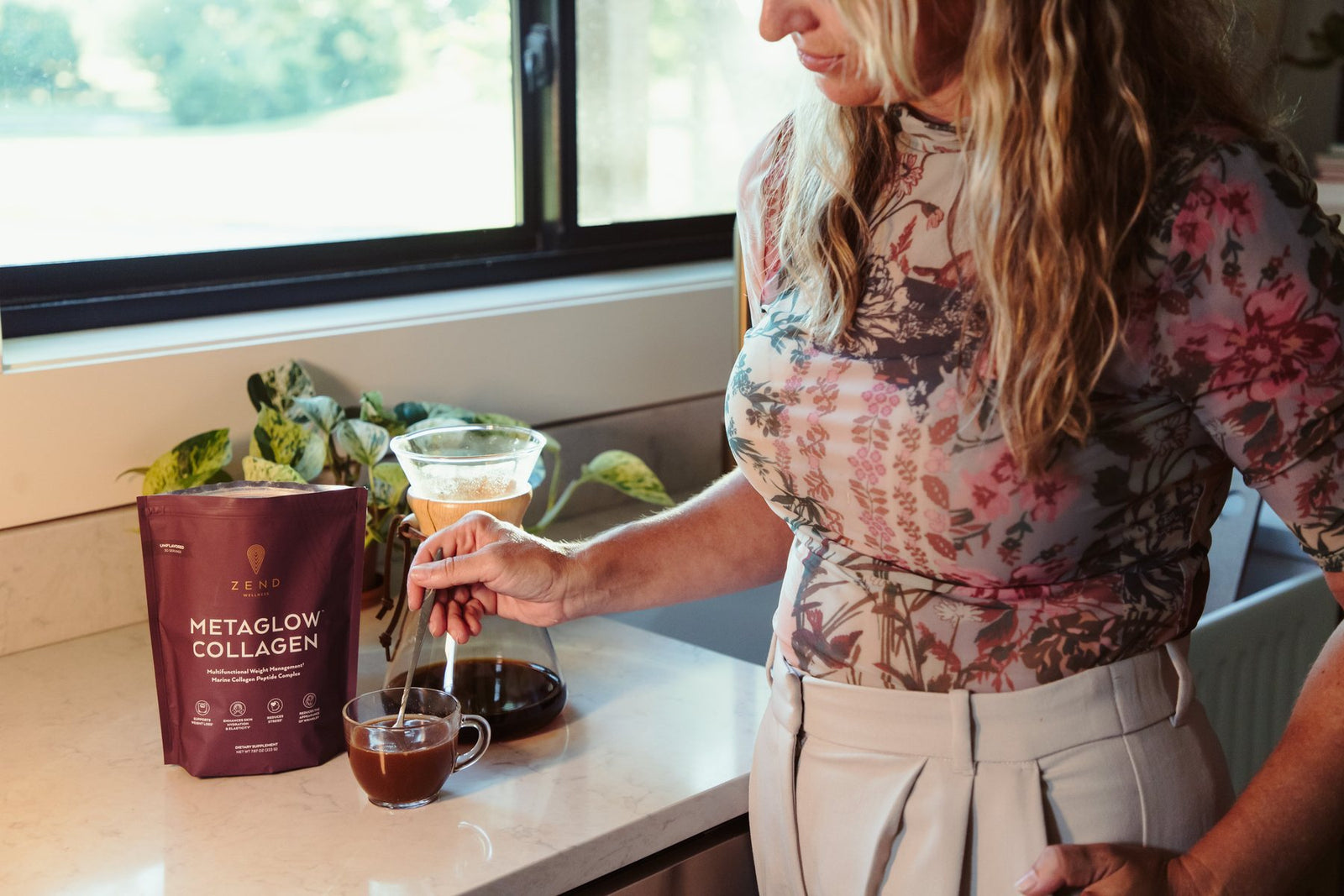 A mother at her kitchen counter, baby on her hip, stirring a scoop of MetaGlow into her coffee